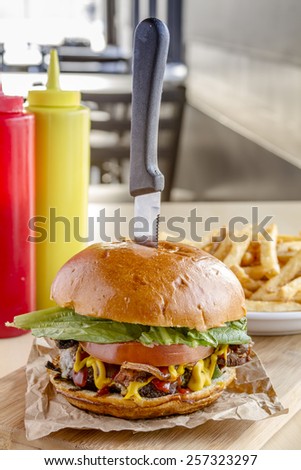 Gourmet pub hamburger with bacon on wooden cutting board with plate of french fries sitting on wooden table