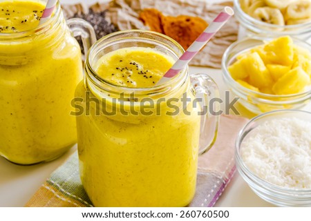 Close up of fresh blended fruit smoothies made with pineapple, banana, coconut, turmeric and chia seeds surrounded by raw ingredients pink straws
