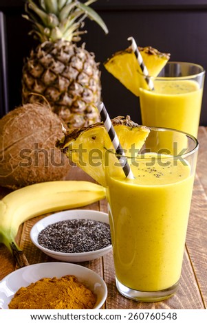 Fresh blended fruit smoothies made with pineapple, banana, coconut, turmeric and chia seeds surrounded by raw ingredients in drinking glass with pineapple slice garnish and blue swirled straw