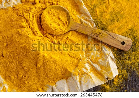 Close up of pile of organic turmeric (curcuma) powder sitting on brown paper with measuring spoon on wooden table