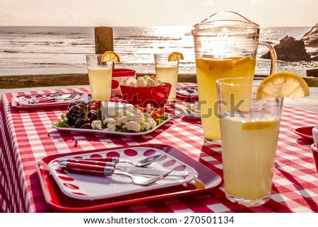 Close up of picnic at the beach overlooking the ocean at sunset with table set with food, dishes, glasses and red checkered table cloth