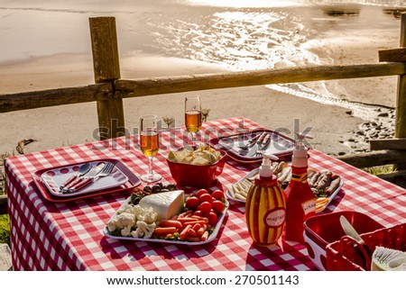 Picnic for 2 at the beach overlooking the ocean with haystack rocks at sunset with table set with food, dishes, wine glasses filled with wine and red checkered table cloth