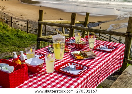 Picnic at the beach overlooking the ocean at sunset with table set with food, dishes, glasses and red checkered table cloth