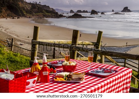 Picnic for 2 at the beach overlooking the ocean with haystack rocks at sunset with table set with food, dishes, wine glasses filled with wine and red checkered table cloth
