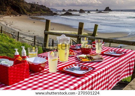 Picnic at the beach overlooking the ocean with haystack rocks at sunset with table set with food, dishes, glasses and red checkered table cloth