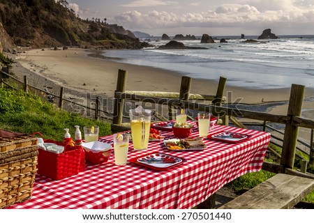 Picnic at the beach overlooking the ocean with haystack rocks at sunset with table set with dishes, glasses and red checkered table cloth