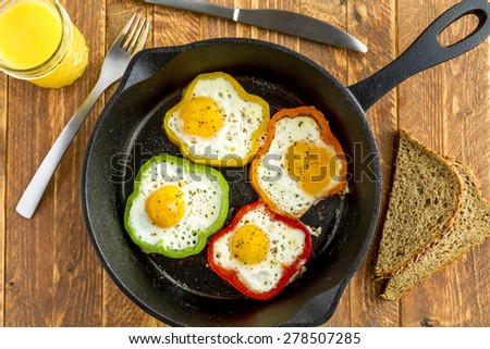 Large cast iron skillet with fried eggs in green, yellow, red and orange bell peppers sitting on wooden table with glass of orange juice and whole wheat toast