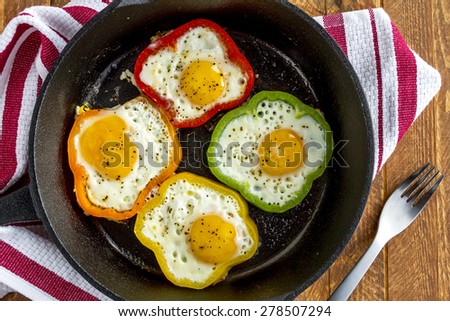 Close up of large cast iron skillet with fried eggs in green, yellow, red and orange bell peppers sitting on wooden table with fork and red striped towel