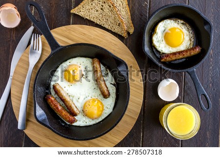 Fried eggs and sausage links in cast iron skillet sitting on kitchen table with whole wheat toast and orange juice