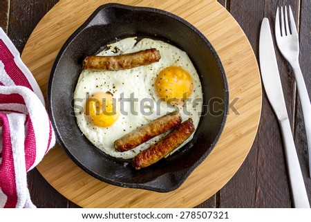Fried eggs and sausage links in cast iron skillet sitting on kitchen table with red striped napkin