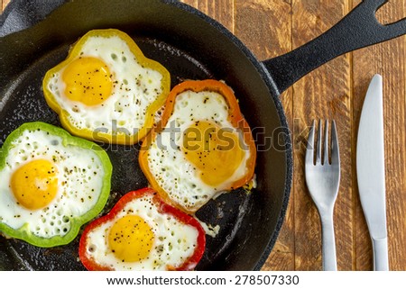 Close up of large cast iron skillet with fried eggs in green, yellow, red and orange bell peppers sitting on wooden table with fork and knife