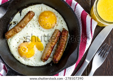 Close up of fried eggs and sausage links in cast iron skillet sitting on kitchen table with orange juice and red striped napkin
