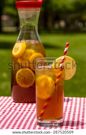 A tall glass and pitcher filled with fresh brewed iced tea and lemon slices with red swirl straws sitting on a red gingham checked tablecloth on picnic table