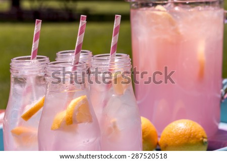 Close up of 4 small glass bottles and pitcher filled with fresh squeezed pink lemonade with pink swirled straws and lemon slices sitting on weather blue drink tray