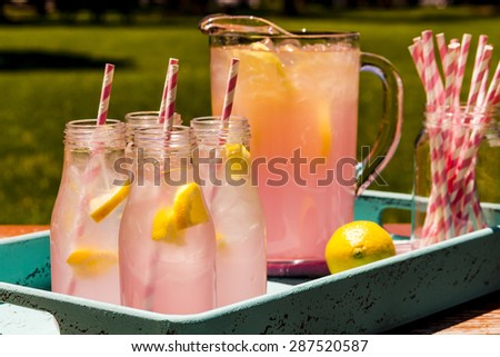 4 small glass bottles and pitcher filled with fresh squeezed pink lemonade with pink swirled straws and lemon slices sitting on weather blue drink tray