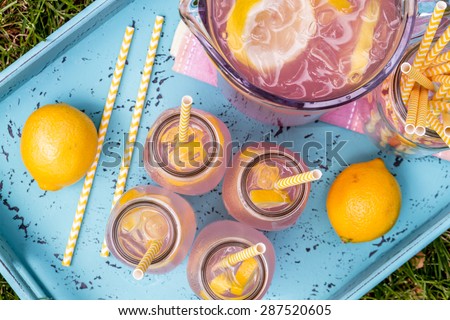 4 small glass bottles and pitcher filled with fresh squeezed pink lemonade with yellow swirled straws and lemon slices sitting on weather blue drink tray from above