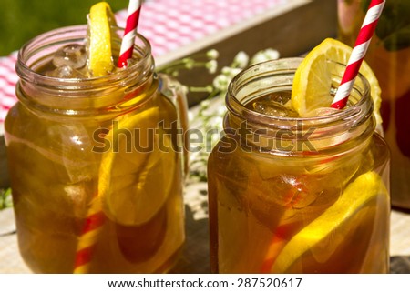 2 mason jar mug and tall pitcher filled with fresh brewed iced tea and lemon slices with red swirl straws sitting in a wooden drink tray on red gingham checked tablecloth on picnic table