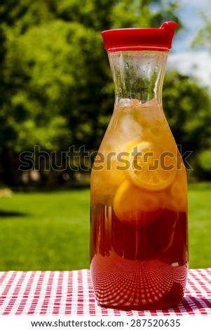 Tall pitcher filled with fresh brewed iced tea and lemon slices with red swirl straws sitting on a red gingham checked tablecloth on picnic table