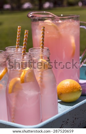 Close up of 4 small glass bottles and pitcher filled with fresh squeezed pink lemonade with yellow swirled straws and lemon slices sitting on weather blue drink tray
