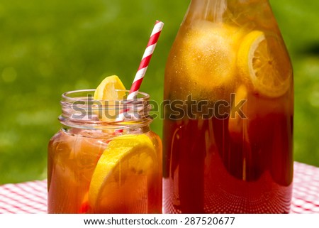 Close up of mason jar mug and tall pitcher filled with fresh brewed iced tea and lemon slices with red swirl straws sitting on a red gingham checked tablecloth on picnic table