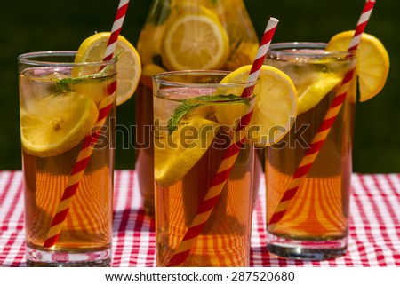 Close up of 3 tall glasses and pitcher filled with fresh brewed iced tea and lemon slices with red swirl straws sitting on a red gingham checked tablecloth on picnic table