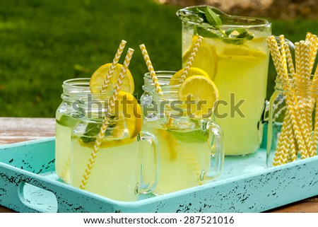 Fresh squeezed lemonade in mason jar mugs and glass pitcher sitting on blue weathered drink tray with yellow straws and fresh lemon slices