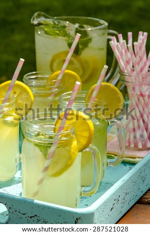 Fresh squeezed lemonade in mason jar mugs and glass pitcher sitting on blue weathered drink tray with pink straws and fresh lemon slices