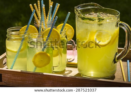 Close up of fresh squeezed lemonade in mason jar mugs and glass pitcher sitting on weathered wooden drink tray with blue and yellow straws and fresh lemon slices
