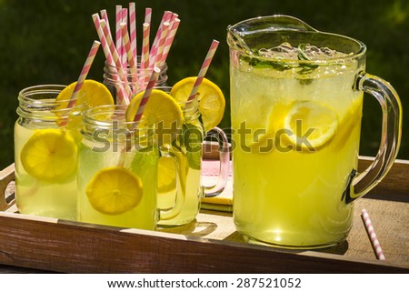 Close up of fresh squeezed lemonade in mason jar mugs and glass pitcher sitting on weathered wooden drink tray with pink straws and fresh lemon slices