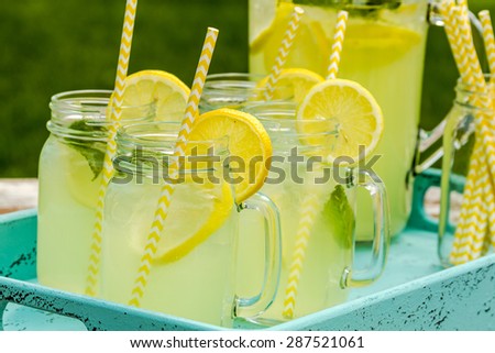 Close up of fresh squeezed lemonade in mason jar mugs and glass pitcher sitting on blue weathered drink tray with yellow straws and fresh lemon slices