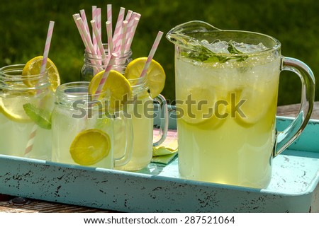 Close up of fresh squeezed lemonade in mason jar mugs and glass pitcher sitting on blue weathered drink tray with pink straws and fresh lemon slices