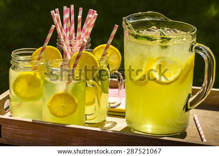 Close up of fresh squeezed lemonade in mason jar mugs and glass pitcher sitting on weathered wooden drink tray with pink straws and fresh lemon slices