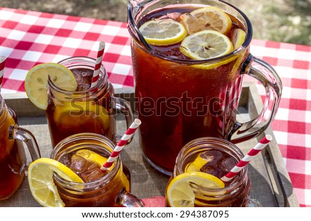 Pitcher and mason jar mugs filled with iced tea and lemons sitting on picnic table with red checked tablecloth