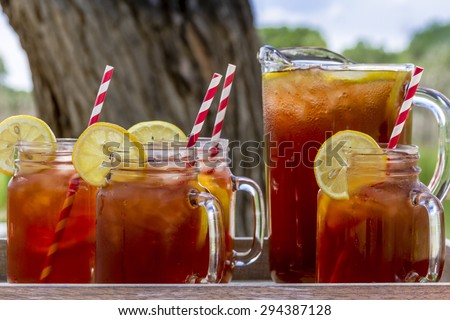Pitcher and mason jar mugs filled with iced tea and lemons sitting on picnic table with red checked tablecloth