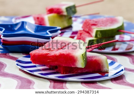 Fresh watermelon slices on a stick sitting on striped plates on picnic table at 4th of July celebration