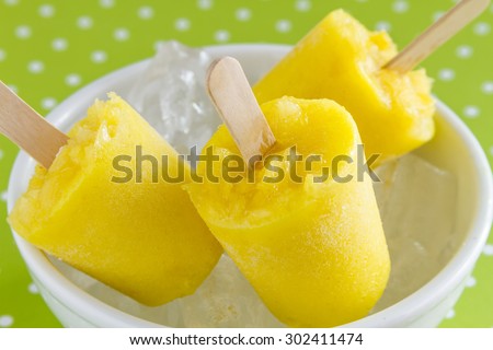 Homemade fresh pureed frozen pineapple popsicles in white bowl with ice sitting on green polka dot background