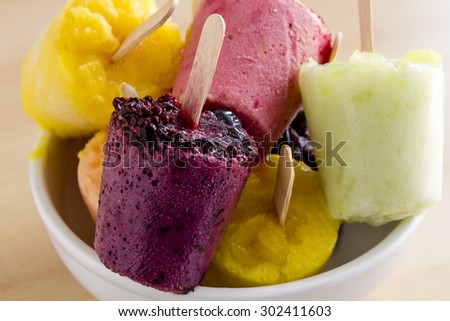 Assorted flavors of homemade fresh pureed frozen fruit popsicles sitting on white bowl on wood table