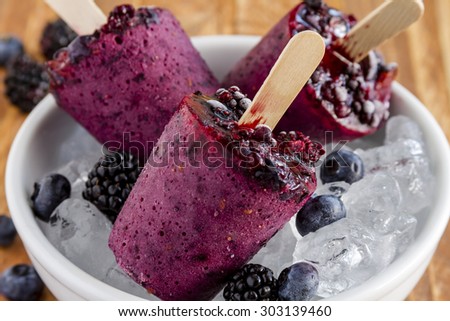 Homemade fresh pureed frozen blueberry and blackberry popsicles in white bowl with ice sitting on wooden table with fresh berries