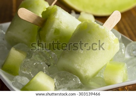 Close up of homemade fresh pureed frozen honey dew melon popsicles on white plate with ice sitting on wooden table with fresh melon pieces