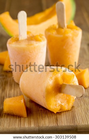 Homemade fresh pureed frozen cantaloupe melon popsicles sitting on wooden cutting board with fresh melon pieces