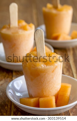 Homemade fresh pureed frozen cantaloupe melon popsicles sitting in small white plates on wooden table with fresh melon pieces