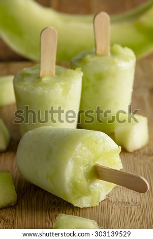 Homemade fresh pureed frozen honey dew melon popsicles sitting on wooden cutting board with fresh melon pieces