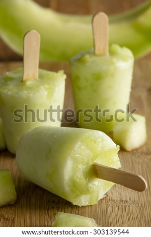 Homemade fresh pureed frozen honey dew melon popsicles sitting on wooden cutting board with fresh melon pieces