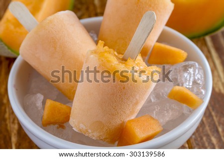 Homemade fresh pureed frozen cantaloupe melon popsicles in white bowl with ice sitting on wooden table with fresh melon