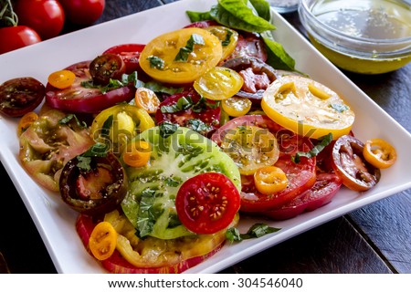 Large square white plate with slices of vine ripe heirloom tomato varieties with fresh basil, salt, pepper, olive oil and fresh herbs