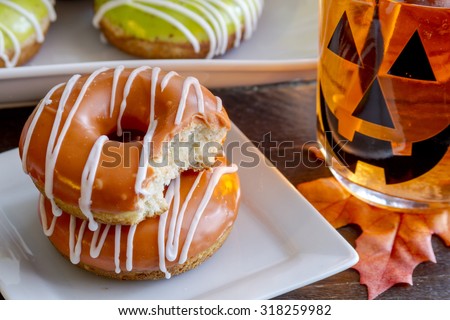 Close up stack of homemade baked pumpkin donuts with orange pumpkin glaze with bite in top donut sitting on white square plate with jack o\' lantern mason jar mug filled with orange drink