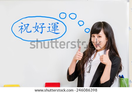 Chinese woman standing by whiteboard showing thumbs up with both hands, with a speech bubble with good luck in Traditional Chinese written inside.