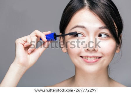 Chinese woman putting mascara on eyelashes on gray background
