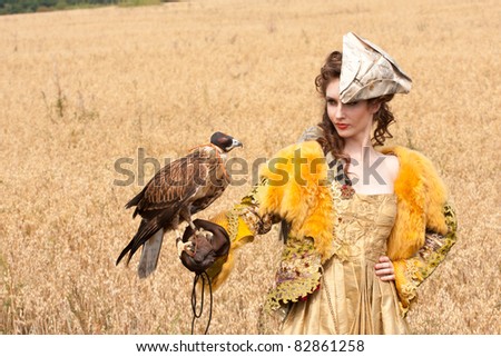 The woman in a beautiful old style dress with falcon has a rest before hunting in yellow field.