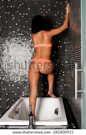 Brunette girl standing in bathtub with his hands on the wall with her back to the photographer
